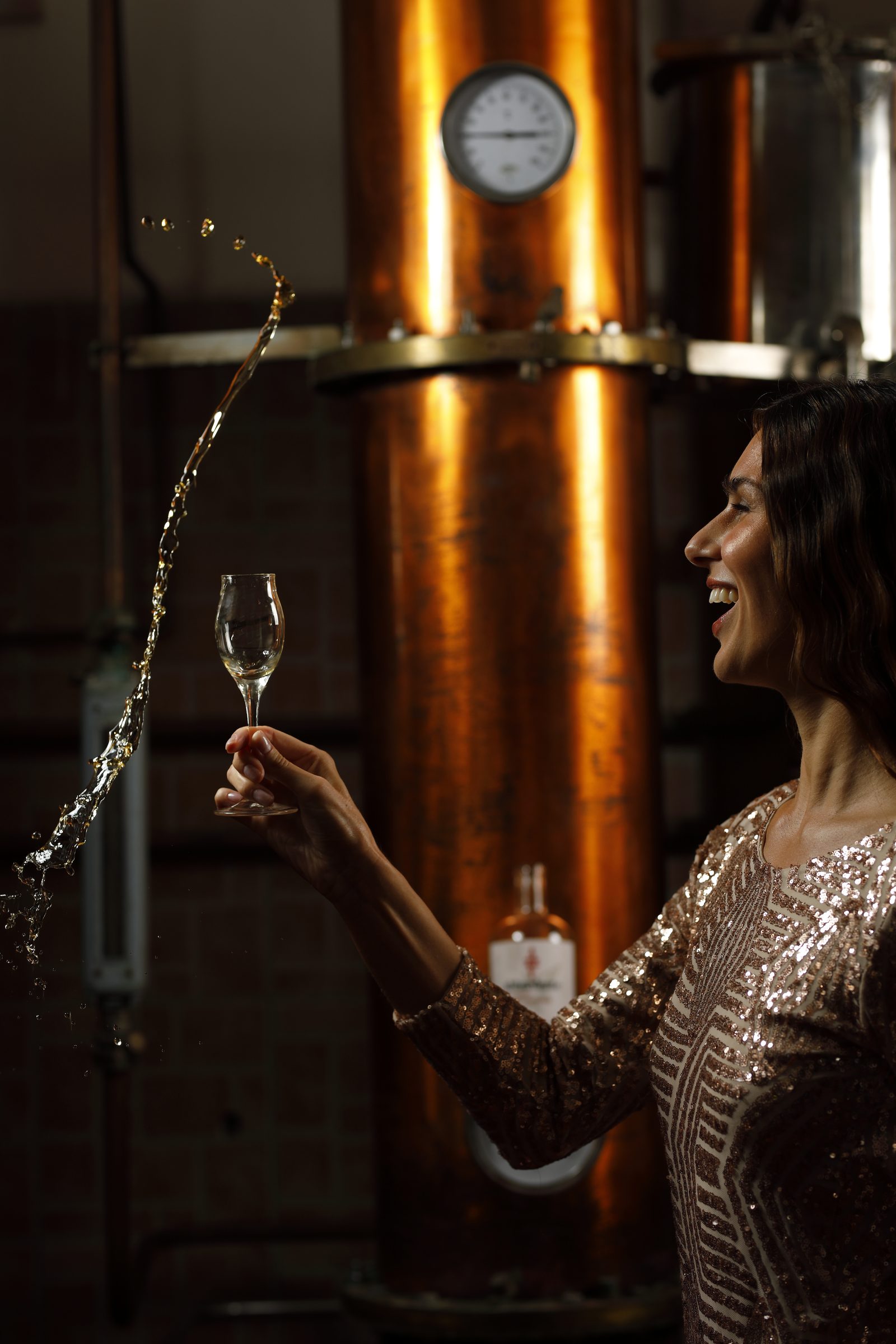 Un calice di distillato sollevato davanti all'alambicco, con uno spruzzo di acquavite in aria
