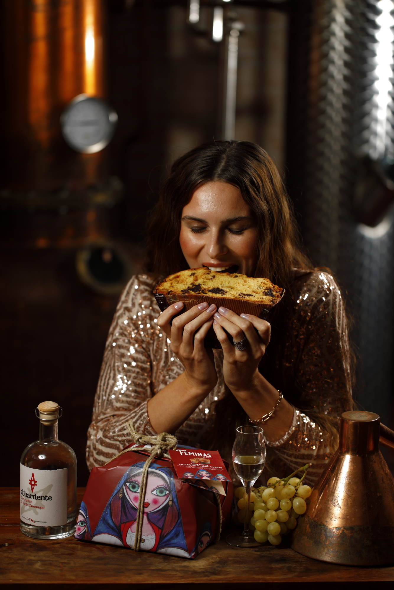 Fetta di panettone farcito portata verso il viso, con la bottiglia di abbardente e il calice sul tavolo di legno